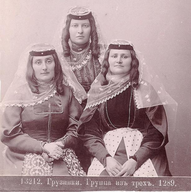 Drei Frauen in traditionellen russischen Hochzeitskleidern mit farbenfroher Stickerei und Perlen, die vor einer einfarbigen weißen Wand in einem Schwarz-Weiß-Foto posieren.