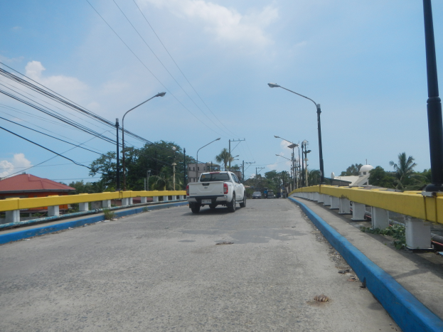 Ein Lastwagen fährt über eine Brücke mit gelben Geländern, Laternenmasten, Strommasten mit Drähten, Bäumen, Gebäuden und einem bewölkten Himmel im Hintergrund.