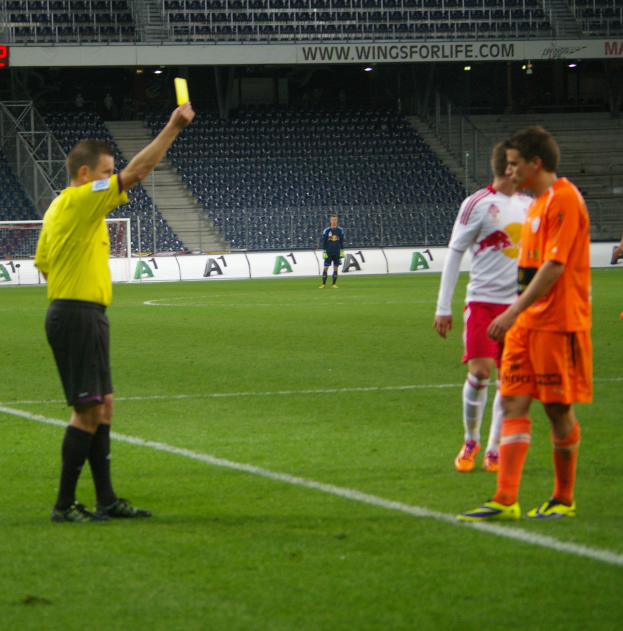 Ein Schiedsrichter zeigt einem Fußballspieler auf dem Feld eine gelbe Karte, umgeben von einer Gruppe von Menschen, mit einem Torpfosten, einem Zaun, einem Schild, Metallrahmen, einem Anzeigebildschirm, Stühlen und einer Decke mit Deckenleuchten im Hintergrund.