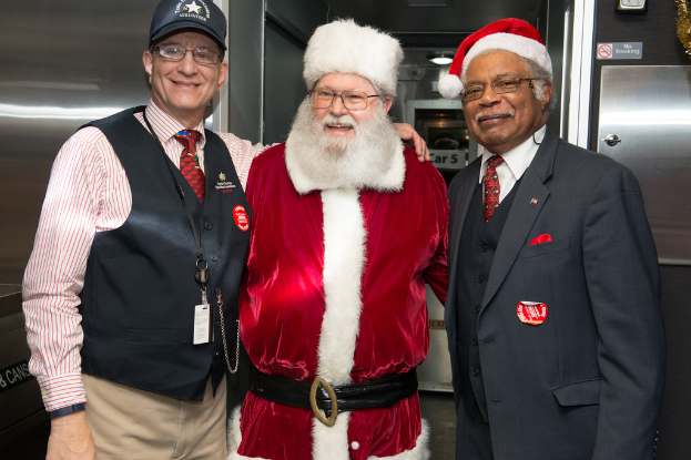 Eine Gruppe von Menschen mit Brillen, Mützen, Krawatten, ID-Karten und Ausweisen, wobei eine Person ein Weihnachtskostüm trägt, vor einer Stahlwand mit Lichtern, Schildern und anderen Gegenständen im Hintergrund.