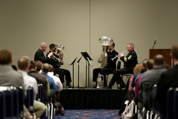 Eine Gruppe von Männern, die auf einer Bühne Blasinstrumente spielen, mit Notenständern, einem Podium mit Mikrofon und einem Publikum, das auf Stühlen sitzt.