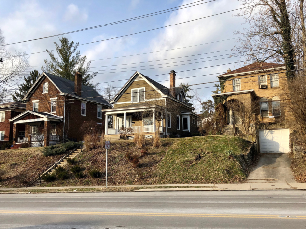 Reihe von Häusern mit Fenstern, Türen, Säulen, Geländern, Stufen, Pflanzen und einem Pfosten mit einer Tafel entlang einer Straße, vor einem bewölkten Himmel.