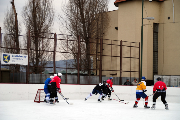 Personen beim Eishockey auf einer Eisfläche mit Gebäuden, Bäumen, einer Straßenlaterne, einem Namensschild und Zäunen im Hintergrund unter einem Himmel.