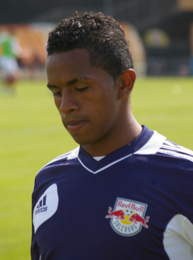 Junger Mann in blauem T-Shirt auf einem Fußballfeld mit unscharfem Hintergrund, der als Flügelspieler mit kurzen dunklen Haaren und entschlossener Miene erscheint.