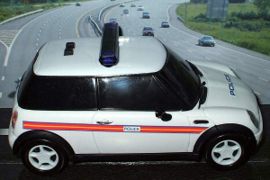 Weißer Polizeiwagen mit "Polizei"-Aufschrift auf einer Straße außerhalb der Stadt, umgeben von fließendem Verkehr, Gras und Bäumen.