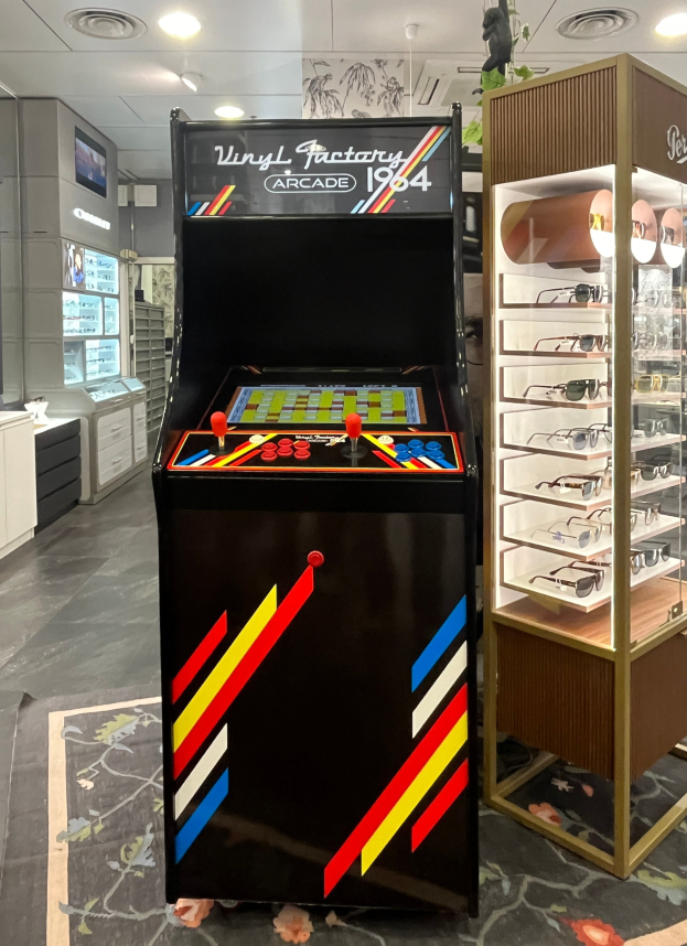 Ein Geschäft mit einem vintage Arcade-Automaten in der Mitte, einem Glasvitrinenschrank auf der rechten Seite und Retro-Stil-Beleuchtung mit Schränken, einem Fernseher und anderen Gegenständen im Hintergrund.