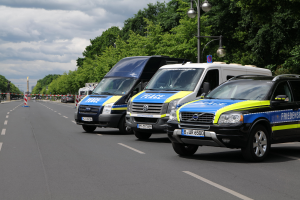 Drei Polizeiwagen auf der Seite einer Straße geparkt, umgeben von Bäumen, Laternenmasten und einem Turm im Hintergrund unter einem bewölkten Himmel.