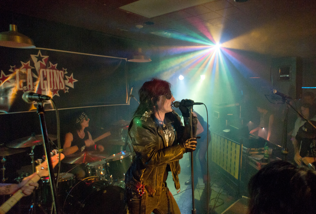 Eine Frau in einer schwarzen Jacke singt in ein Mikrofon auf der Bühne, im Hintergrund ein Mann mit Sonnenbrille und Stirnbinde, der eine Bassgitarre spielt; im Hintergrund ist ein "LA Guns"-Plakat und Diskoleuchten zu sehen.
