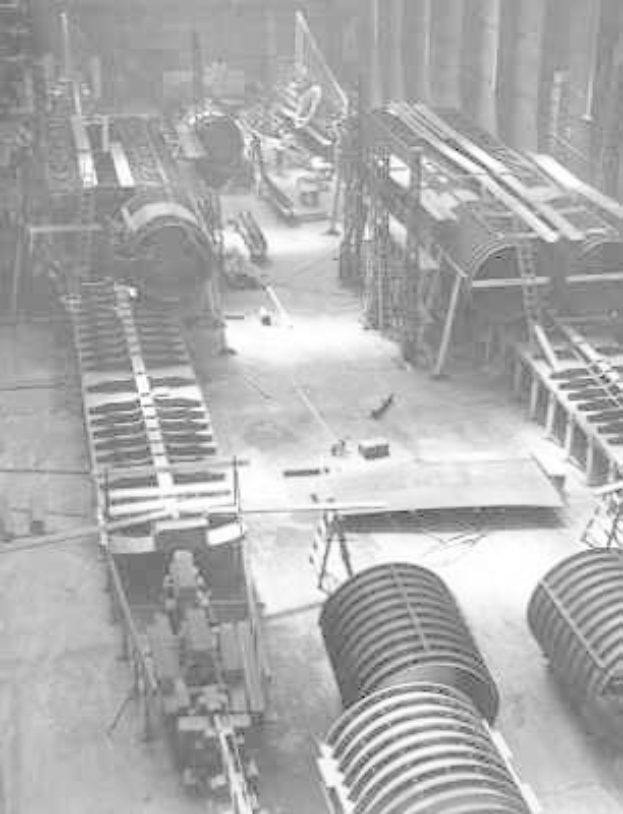 Schwarz-weiß-Foto eines industriellen Fabrikinneren mit zahlreichen Rohren, Maschinen und Geräten auf dem Boden, vor strukturellen Säulen und einer Wand.