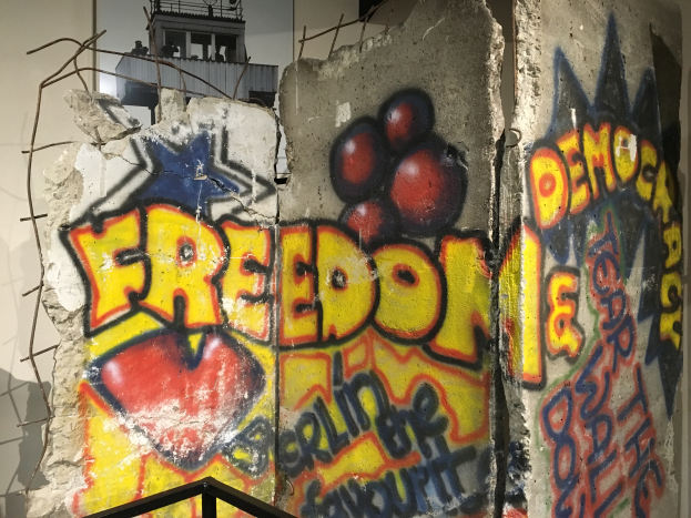 Eine Wand mit mutiger, schwarzer Graffiti, die "Freiheit und Demokratie" lautet, in einem Raum mit einem Geländer unten und einem Gebäude mit Fenstern im Hintergrund.