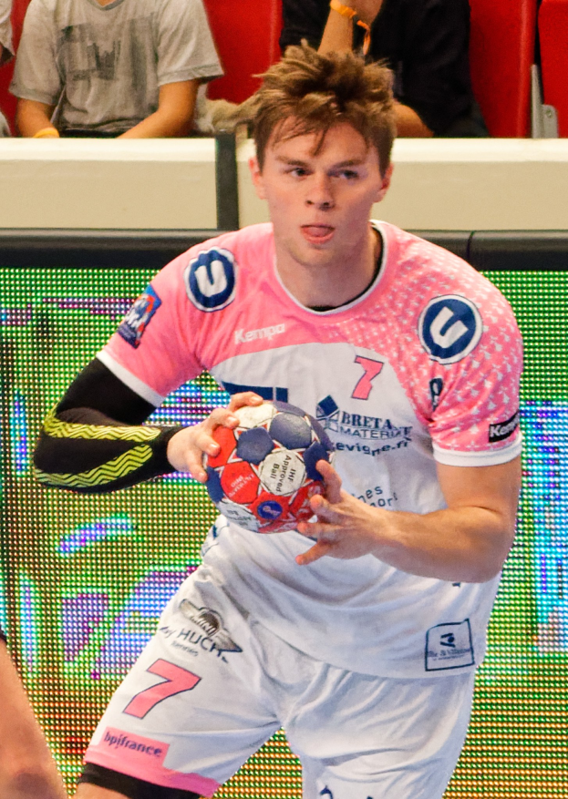 Ein Mann in einem pinken Hemd, der einen Fußball hält, mit einer anderen Person Hand auf der linken Seite und Menschen im Hintergrund auf Stühlen sitzend.