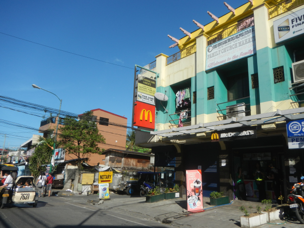 Eine Straßenszene mit einem McDonald's-Restaurant an der Ecke, umgeben von Gebäuden, Bäumen, Straßenlaternen, Fahrzeugen, Fußgängern und Werbetafeln unter einem klaren blauen Himmel.