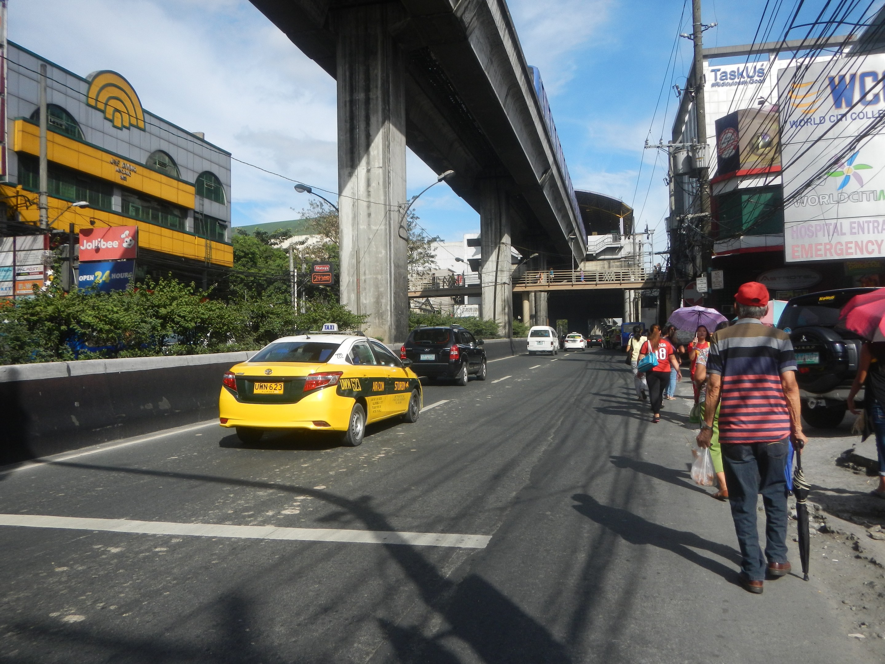 Ein gelbes Taxi fährt auf einer Stadtstraße mit hohen Gebäuden, Passanten mit Regenschirmen, einer Brücke im Hintergrund und bewölktem Himmel.
