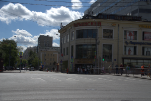 Eine Stadtstraße mit Menschen, hohen Gebäuden, Schildern, Ampeln, einem Metallzaun, Fahrzeugen, Bäumen und einer bewölkten Himmel.
