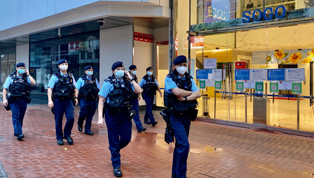 Eine Gruppe von Polizisten in Gesichtsmasken, die vor einem Gebäude mit Glaswänden und -türen die Straße entlanggehen, mit einem Geschäft auf der rechten Seite, das Plakate auf seinen Glaswänden zeigt.