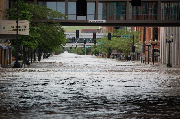 Überschwemmte Stadtstraße mit Wasser, das die Straße, Masten, Laternen, Verkehrsschilder, Bäume, Gebäude und eine Brücke im Hintergrund bedeckt.
