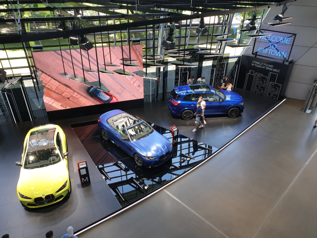 Eine Gruppe von Menschen steht im BMW Museum in München, Deutschland, mit Autos auf der linken Seite und einem Schild mit der Aufschrift "BMW Museum München" auf der rechten Seite, mit Blick auf eine städtische Außenansicht durch Glasfenster.