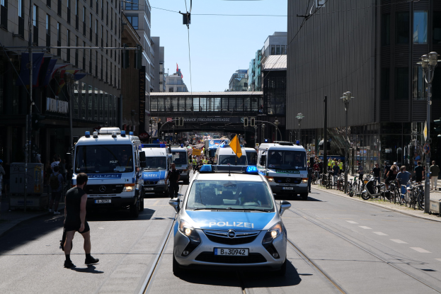 Polizeifahrzeuge fahren eine Stadtstraße mit hohen Gebäuden, Fußgängern auf dem Bürgersteig, geparkten Fahrrädern, Laternen, einer Brücke im Hintergrund und einem klaren blauen Himmel.