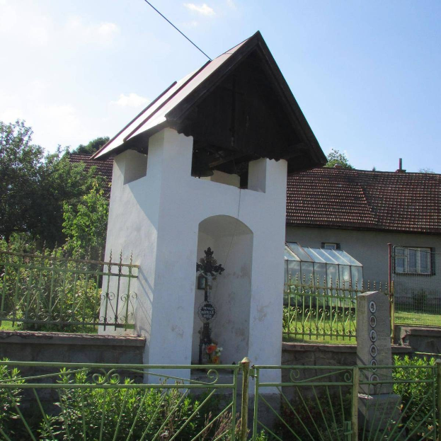 Kleine weiße Kirche mit einem Kreuz darauf, umgeben von einem Metallzaun, Pflanzen und einem Weg, vor Häusern, Pfählen, Drähten und einem bewölkten Himmel.