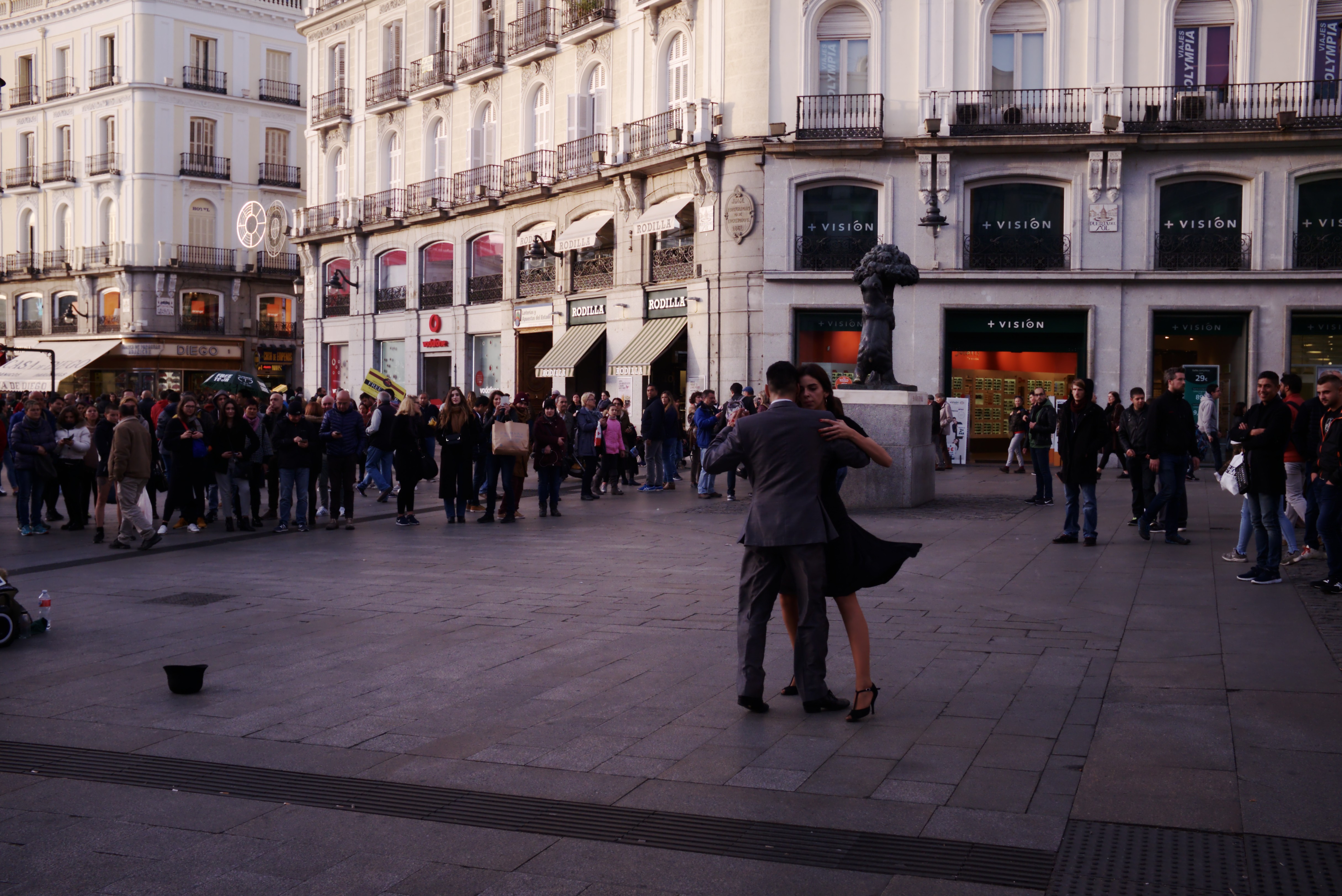 Ein Mann und eine Frau tanzen auf einem belebten Stadtplatz, umgeben von Zuschauern, mit einem Kinderwagen, Eimer und Stangen links und Gebäuden, Gelöndern, Schildern, einer Statue und einem bewölktem Himmel im Hintergrund.