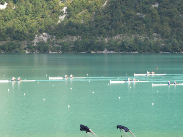 Eine Gruppe von Menschen, die in einem Wettbewerb auf einem Gewässer mit Bäumen und einem Hügel im Hintergrund rudern, und Kleidung über Stühle am Boden drapiert.