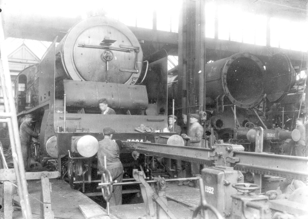 Schwarzes und weißes Foto von Männern, die an einem Zug in einer Fabrik arbeiten, einige halten Werkzeuge, mit einer Leiter auf der linken Seite und einer Hütte im Hintergrund.