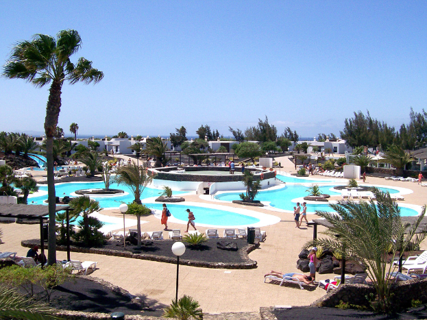 Ein Resort mit Pools umgeben von Bäumen, Menschen die dazwischen gehen, Gebäude im Hintergrund und ein klarer Himmel darüber.