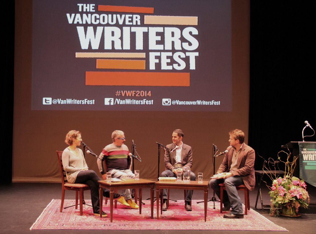 Eine Gruppe von Männern und eine Frau sitzen auf Stühlen auf einer Bühne, sprechen in Mikrofone, mit einem Bildschirm im Hintergrund, einem Teppich auf dem Boden, einem Tisch in der Mitte und einem Blumentopf neben ihnen.