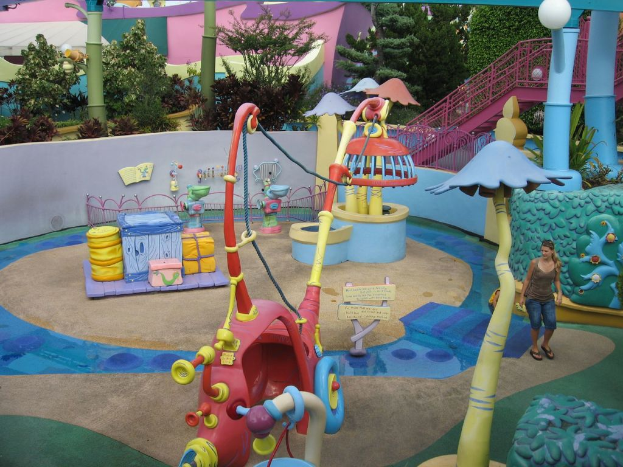 Eine Frau steht in der Nähe von Spielgeräten für Kinder, mit Säulen, einem Metallzaun, Bäumen und einem Haus im Hintergrund.