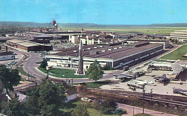 Alte Schwarz-Weiß-Airport-Szene mit geparkten Propellerflugzeugen, Terminalgebäuden, Bäumen, Straßen, Fahrzeugen, Strommasten, Gras, Bahnschienen, fernen Türmen, rollenden Hügeln und einem klaren Himmel; Bildunterschrift unten.