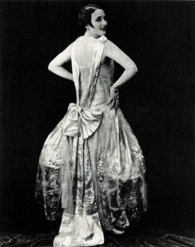 Eine Frau in einem Kleid im Stil der 1920er Jahre mit hohem Kragen, langen Ärmeln und einem voluminösen Rock steht selbstbewusst mit einer eleganten Hochsteckfrisur und dezentem Make-up.