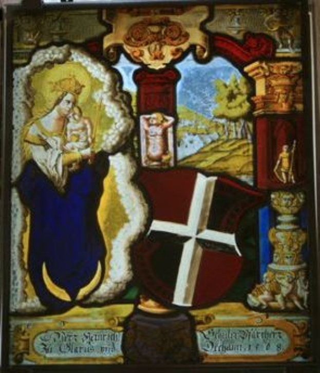 Ein Buntglasfenster in einer Kirche, das die Jungfrau Maria in einem weißen Kleid und einem Heiligenschein zeigt, die ein Baby hält, umgeben von einem Kreuz, einem Schild, einer Krone und religiöser Schrift in einer Schrifttype.