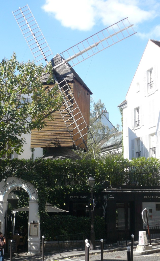 Eine Windmühle, mehrere Gebäude, Bäume, Metallstangen und ein Zaun in einer ländlichen Landschaft.