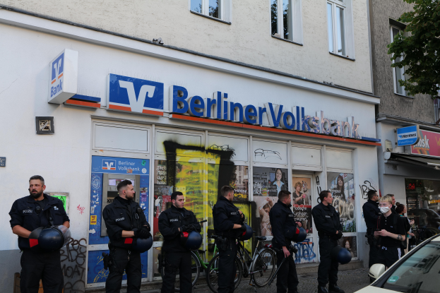 Eine Gruppe von Polizisten steht vor einer Berliner Volksbank, einige halten Helme, mit einem Auto rechts, einem Baum links und Gebäuden mit Fenstern und Texttafeln im Hintergrund.