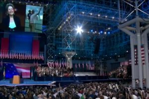 Eine große Menge sitzt vor einer Bühne mit einem Podium, Mikrofon, Flaggen, einem Bildschirm und Lichtausrüstung und wartet auf eine Präsidentschaftsdiskussion.