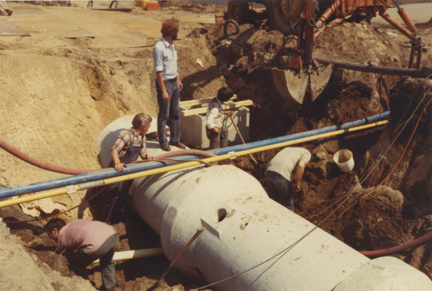 Eine Gruppe von Männern arbeitet an einem großen Rohr im Schmutz, umgeben von verschiedenen Werkzeugen und Geräten, mit einem Fahrzeug und einer Straße im Hintergrund.