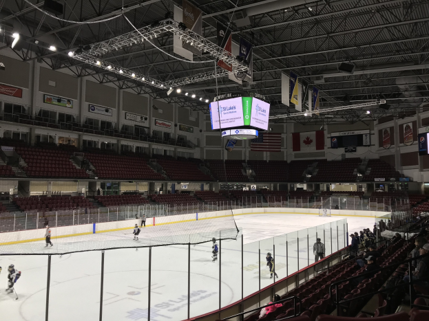 Eishockeyspiel in einer großen Arena mit einem zentralen Netz, Zuschauern auf beiden Seiten, Deckenleuchten und Trägern sowie Bannern an den Wänden, die "St. Louis Blues Arena" beschriftet sind.