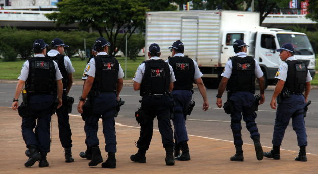 Eine Gruppe von Beamten in Uniform, einschließlich Mützen, Hemden, Schutzwesten, Uhren, Hosen und Schuhen, die auf einer Straße mit einem geparkten Lastwagen auf der rechten Seite neben einem Zaun gehen. Im Hintergrund sind Gebäude, Bäume, Pflanzen, Pfosten und Schilder zu sehen, wobei in der oberen rechten Ecke eine Brücke zu erkennen ist.