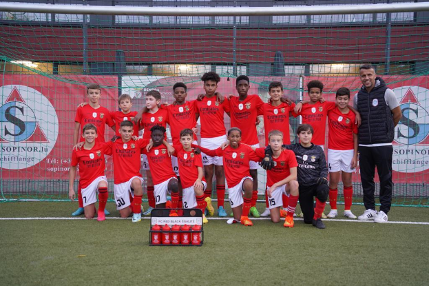 Eine Gruppe junger Männer in roten T-Shirts und schwarzen Shorts steht auf einem Fußballfeld neben einer Kiste roter Gegenstände, im Hintergrund Schilder mit der Aufschrift "FC Rot-Schwarz Eglaute - Champions League".