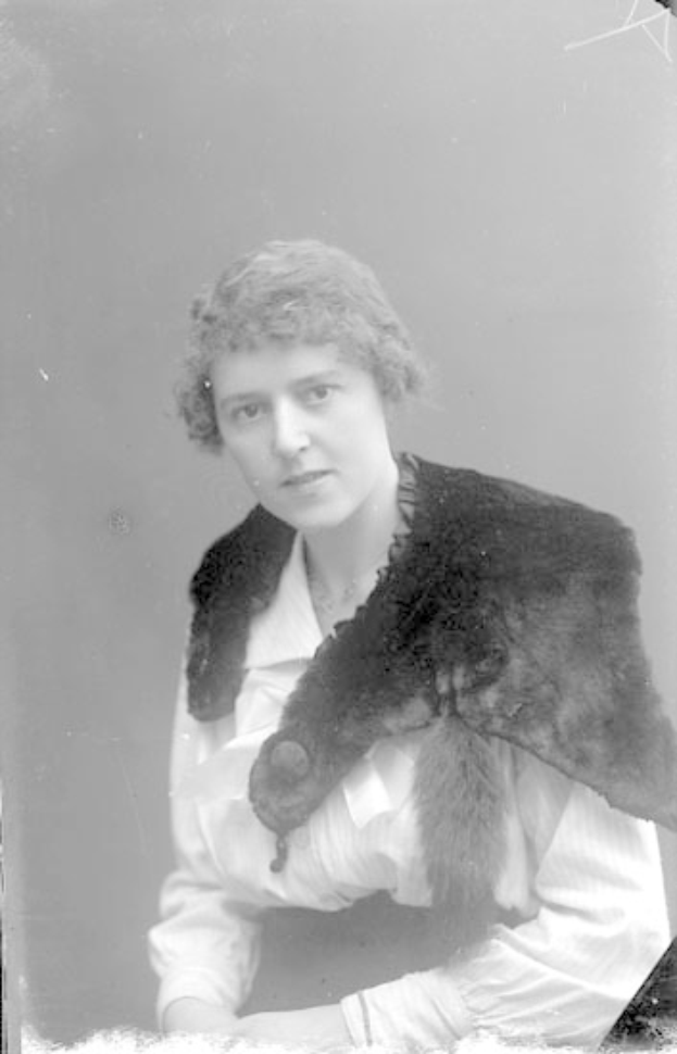 Eine Frau in einem Schwarz-Weiß-Foto trägt einen Pelzmantel, ihr Haar ist hochgesteckt und sie hat einen ernsten Gesichtsausdruck, während sie in die Ferne schaut und ihre Hände gefaltet hat.