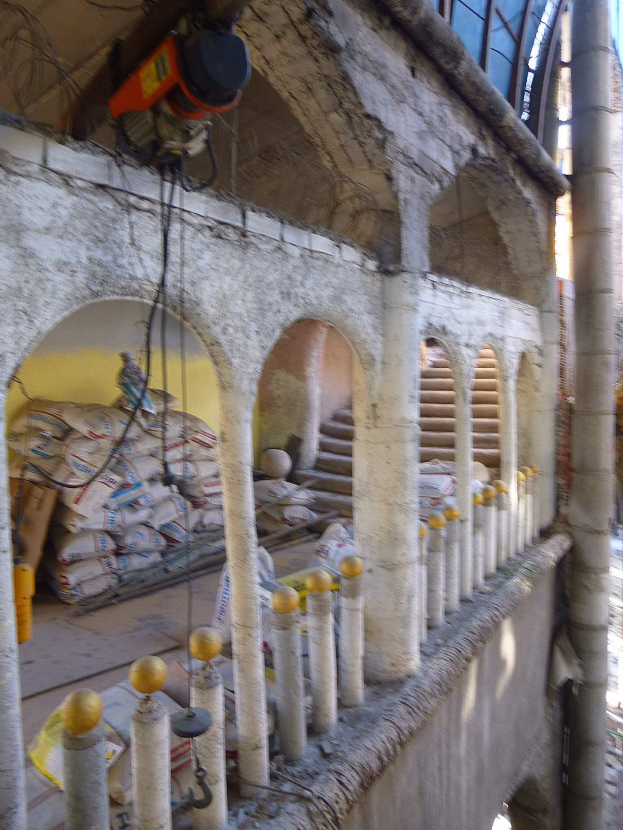 Altes Gebäude mit Bogengängen, Säulen und Treppen wird renoviert, mit verstreuten Werkzeugen, Materialien und Gegenständen, darunter Taschen und eine Maschine mit Drähten oben drauf.