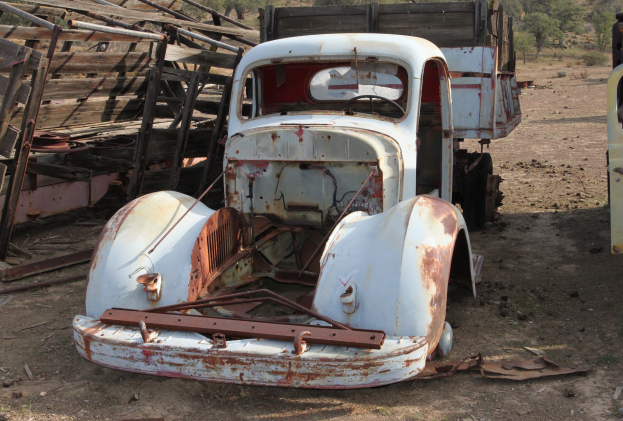 Ein alter, rostiger Lastwagen in einem Zustand der Verwahrlosung sitzt im Dreck, umgeben von einem Holzzaun mit Bäumen im Hintergrund.