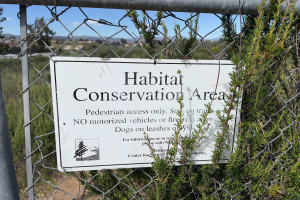 Ein Schild an einem Kettenzaun mit der Aufschrift "Habitat-Schutzgebiet" vor Pflanzen, Bäumen, Hügeln und einem klaren blauen Himmel im Hintergrund.