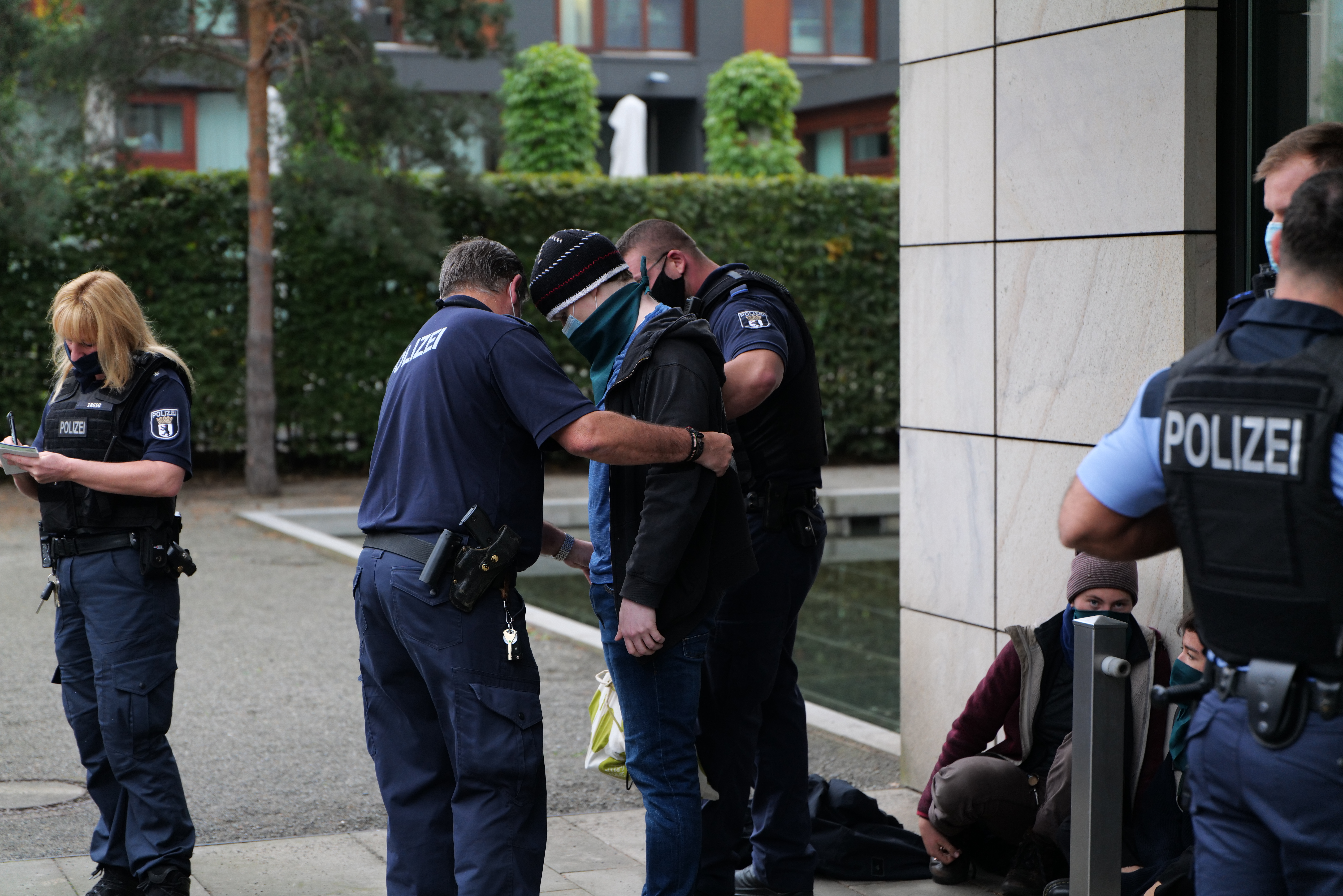 Eine Gruppe von Polizisten steht vor einem Gebäude zusammen, eine Frau hält ein Buch und einen Stift in der Hand, während einige Menschen in der Nähe stehen und sitzen und Pflanzen, Bäume und Gebäude im Hintergrund zu sehen sind.