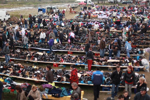 Eine belebte Marktszene mit zahlreichen Menschen, Tischen mit Schuhen, Kleidung und anderen Gegenständen, einer Tasche rechts und Fahrzeugen und Zelten im Hintergrund.