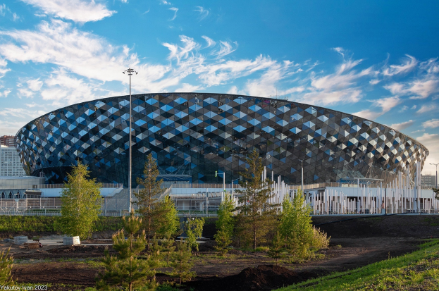 Das Olympische Stadion in Sotschi, Russland, ein großes Gebäude mit einzigartigem Design, umgeben von Bäumen, Pflanzen, Gras, Pfählen und einem Zaun, mit anderen Gebäuden und einem bewölkten Himmel im Hintergrund.