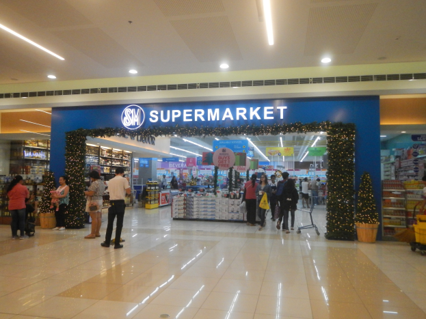 Eine Gruppe von Menschen vor einem Supermarkt, einige mit Taschen, Einkaufswagen, Topfpflanze, Regalen mit Gegenständen, Schildern und einer Decke mit Leuchten.