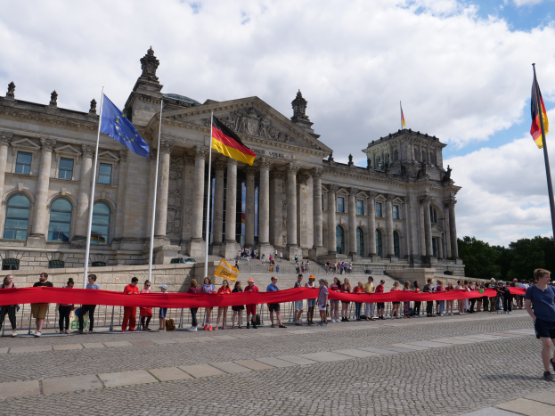 Eine Gruppe von Menschen vor dem Reichstaggebäude in Berlin, Deutschland, das mit Fenstern, Säulen, Bögen und Statuen geschmückt ist und von Fahnen mit Stangen umgeben ist, mit einem roten Band im Vordergrund, einer Person auf der rechten Seite und Bäumen und einem bewölkten Himmel im Hintergrund.