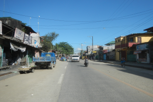 Eine Straße in der Stadt mit Fahrzeugen, einem Radfahrer auf der rechten Seite, Gebäuden auf beiden Seiten, Bäumen im Hintergrund und einem klaren blauen Himmel.