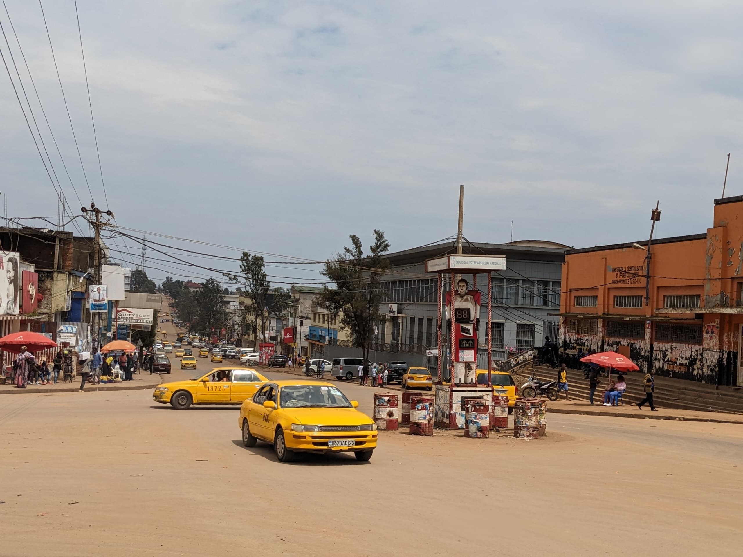 Ein gelbes Taxi fährt auf einer belebten Stadtstraße mit Gebäuden an der Seite, wo Fußgänger mit Schirmen unter einem bewölkten Himmel gehen, mit Strommasten und Drähten im Hintergrund.